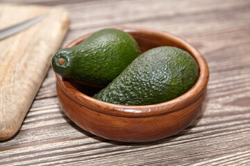 Two ripe whole avocados with textured dark green skin placed in a smooth wooden bowl on a striped rustic wooden table beside a cutting board