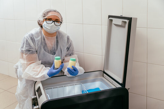 Medical professional in mask and gloves holds two breast milk containers near open medical freezer. Used in donor milk banks and neonatal hospital care units.