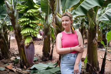 Young Caucasian woman smiling gently and enjoying the nature surrounding her, a banana plantation with lots of healthy trees and bountiful clusters of green bananas in Tenerife, Canary Islands, Spain