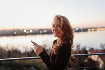 Young woman enjoying sunset by the river while using her smart phone on a balcony in a serene location © Marko