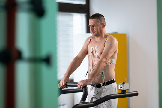 A middle-aged man is undergoing a medical stress test on a treadmill, connected to ECG electrodes for heart monitoring in a clinical environment