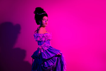 Elegant young woman in a vintage gown posing with a vibrant neon pink backdrop showcasing historical fashion and regal elements