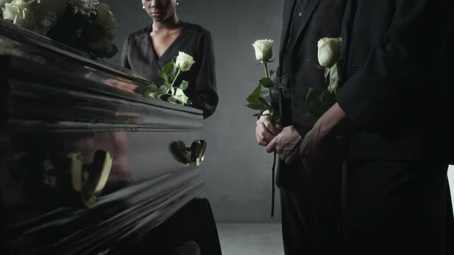 Low angle shot of people in black, holding white roses, standing mournfully beside polished dark coffin adorned with white flowers at funeral