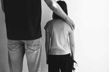 Black and white image of man standing beside a child with his hand gently resting on the child's shoulder, child holding a plush toy, white wall, concept of child abuse, kidnapping violence children