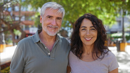 Middle-aged couple smiling together outdoors on a sunny day, capturing love and companionship in a vibrant urban park with lush greenery and contemporary buildings in the background.