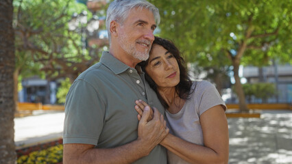 Fototapeta premium Middle-aged couple embracing outdoors in a sunny urban park, expressing love and togetherness, with man and woman standing closely amidst greenery and modern urban landscape.