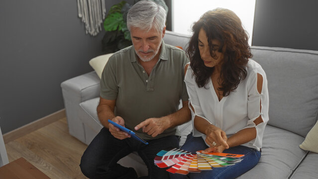 Middle-aged couple in living room choosing paint colors with swatches, man and woman engaged in home decor, sitting together in a bright and cozy indoor space.