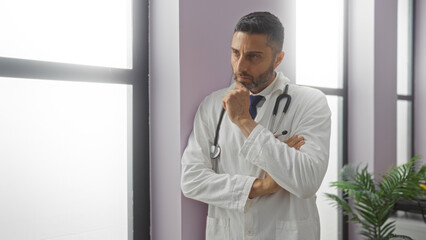Doctor thinking in a clinic room with a stethoscope, reflecting on a case by the window, conveying...