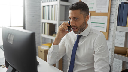 Man talking on phone in an office, wearing a white shirt, with a computer, looking surprised, sitting at a desk, surrounded by shelves, papers, and a corkboard.