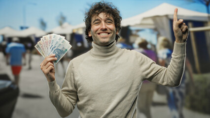 Hispanic man smiling holds polish zloty banknotes pointing upward outdoors on a busy city street.