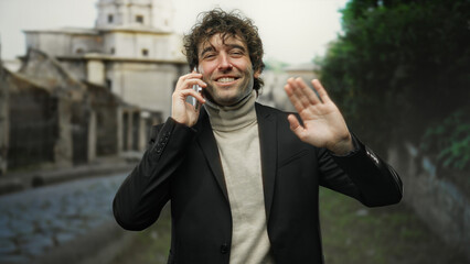 Man smiling outdoors, dressed in a suit, talking on smartphone in city street, showcasing a cheerful demeanor while waving, reflecting a casual urban ambiance.