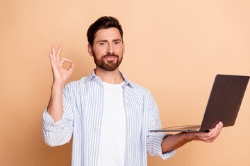 Confident young man holding a laptop making an okay hand gesture against a beige background,...
