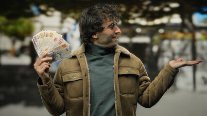 Hispanic man outdoors in a city street extends his hand, holding turkish lira banknotes, creating a vibrant scene of currency and opportunity.