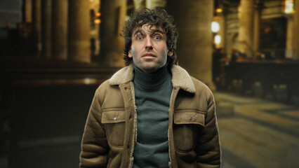 Young man with curly hair stands indoors in a historic church, wearing a warm brown jacket,...