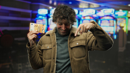 Fototapeta premium Hispanic man indoors at a casino, holding a 50 euro banknote, smiling while pointing down, with blurred slot machines in the background, evoking a sense of winning and fortune.