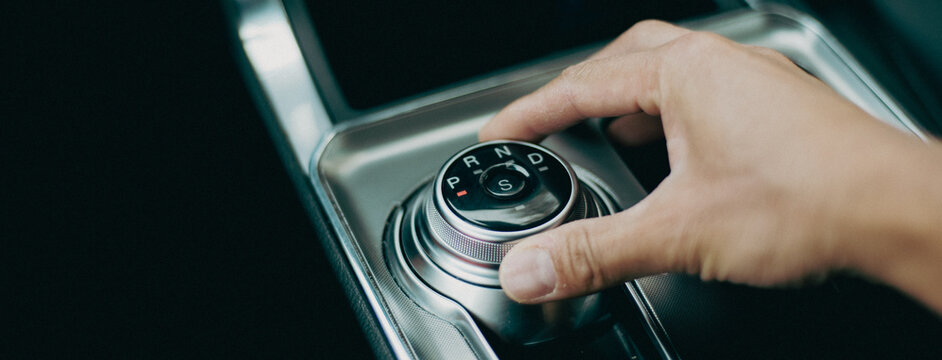Panorama view Asian man hand rotates rotary shifter gear shift knob at middle console of luxury SUV car in America, automatic transmission with dial gear selector of P, R, N, D and S Sport mode