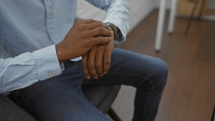 Obraz premium Man checking wristwatch while seated in a modern office environment, showcasing a focused african american professional in a casual blue shirt and jeans.