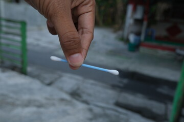 A hand holds a cotton swab.  The swab is light blue and white.  Outdoor setting with blurred background
