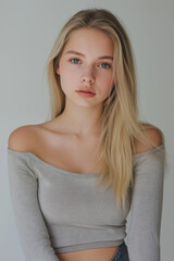 Fototapeta premium Close-up portrait of a young woman with long, straight blonde hair. She is wearing a light grey off-shoulder top. The background is a plain, light grey, providing a neutral backdrop that emphasizes
