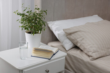 Bedside table with book, houseplant and glass of water near bed in room