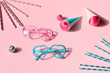 Heart-shaped sunglasses in pink and blue on a pastel pink background with striped straws, candy,...