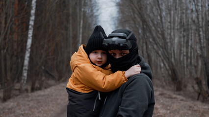 A young man in a mask with FPV glasses on his head, holding his son in his arms,