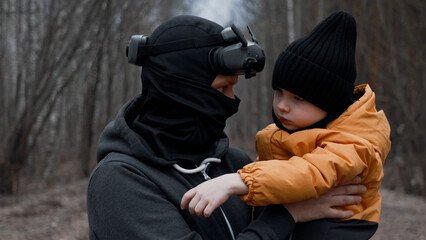 A young man in a mask with FPV glasses on his head, holding his son in his arms,