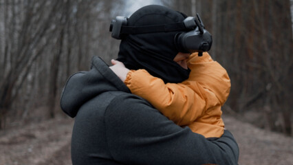 A young man in a mask with FPV glasses on his head, holding his son in his arms,