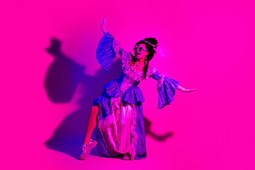 Elegant woman in historic costume posing under neon pink lighting with a playful pose and modern accessories