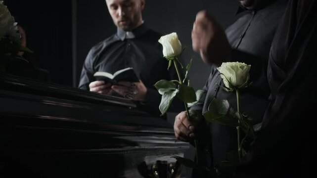Close-up low angle shot of priest in black, holding open book, standing beside mourners with white roses near polished casket at somber, respectful funeral service