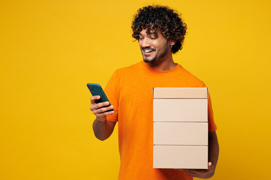 Young fun happy Indian man he wear orange t-shirt casual clothes hold stack cardboard blank boxes use mobile cell phone online isolated on plain yellow background studio portrait. Lifestyle concept.