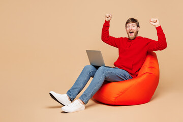 Full body young IT man wear red hoodie casual clothes sit in bag chair hold use work on laptop pc...