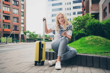 Fototapeta premium Young woman with suitcase looking at phone while sitting on urban street during daylight travel adventure