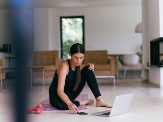 A woman in sportswear is sitting in the living room and preparing for online training while using a...
