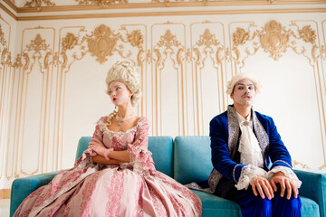 Elegant couple in baroque costumes posing on a vintage sofa in a luxurious palace interior