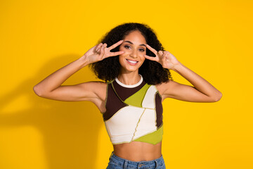 Young woman showing peace signs in casual top against yellow background, cheerful and stylish fashion