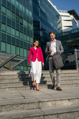 Business people walking and talk to each other in front of modern office. Happy excited office colleagues in stylish outfits talking about work when walking over street in summer