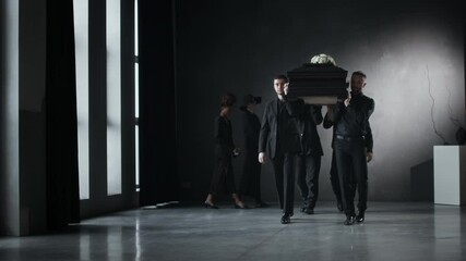 Low angle shot of funeral procession of mourners in black formal wear carrying coffin through a minimalist, dimly lit space with large windows