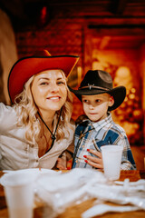 Smiling woman in a red cowboy hat poses with a young boy in a black cowboy hat at a rustic indoor celebration, surrounded by cups and plates on the table..
