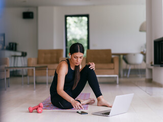 A woman in sportswear is sitting in the living room and preparing for online training while using a...
