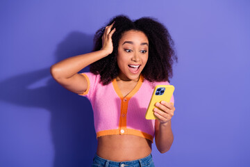 Excited young woman holding a smartphone while smiling in surprise against a vibrant purple...