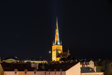 Fototapeta premium Illuminated Oleviste Church rises above the rooftops of Tallinn under Estonia's starry night sky, with the visible moon.