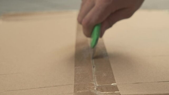 hand cuts through adhesive tape on a cardboard box using a green utility knife. The sharp blade for easy opening.
