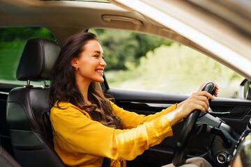 Cheerful young woman driver sitting in front seat, holding auto steering wheel, driving her new car...