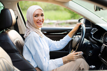 Car insurance concept. Happy young muslim lady sitting in modern vehicle, enjoying driving auto, holding steering wheel and smiling at camera inside luxury salon