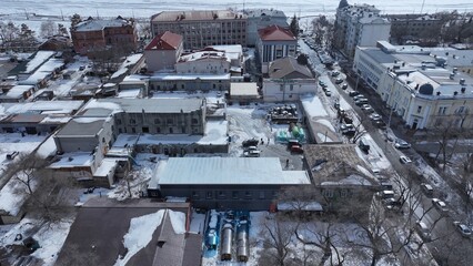 Winter aerial view of a city with a mix of old and modern buildings, snowy streets, and parked cars. Some buildings appear under renovation. Bare trees and snow-covered rooftops highlight the cold.