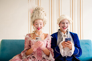 Elegant couple in baroque costumes having fun with smartphones indoors