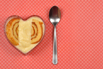 A heart-shaped glass bowl with a roll cake and vanilla cream next to a spoon on a red patterned background, shot from above.