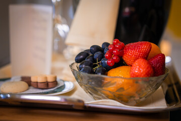 A fruit bowl with strawberries, oranges, red currants, and grapes sits beside a plate of cookies on a wooden surface in a boutique hotel in Cortina, Italy.