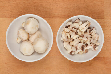 Two bowls with mushrooms, one with whole and the other with sliced mushrooms, on a wooden surface, shot from above